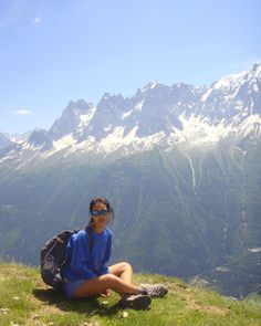 a woman sitting on top of a lush green hillside next to a mountain range with snow covered mountains in the background