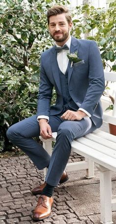 a man in a blue suit sitting on a white bench wearing brown shoes and a boutonniere