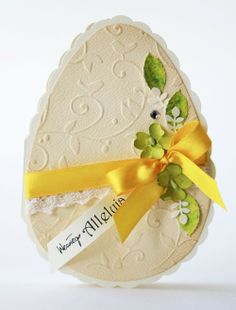 an egg shaped card with yellow ribbon and flowers on the front, sitting on a white surface