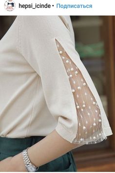 a woman wearing a white shirt and green pants with pearls on the sleeves is looking at her watch