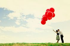 a man and woman holding red balloons in their hands while standing on a hill under a cloudy blue sky