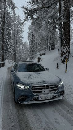 a car driving down a snow covered road in the middle of some trees and bushes