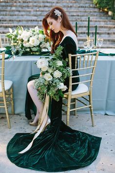 a woman sitting at a table with flowers and greenery in her hair, dressed in an emerald green velvet gown