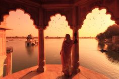 a woman standing on top of a balcony next to a body of water at sunset