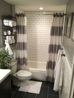 a white toilet sitting next to a bath tub under a bathroom window covered in gray and white striped curtains