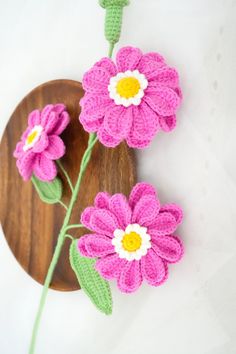 two crocheted flowers on a wooden plate with green stems in the middle and one pink flower at the end