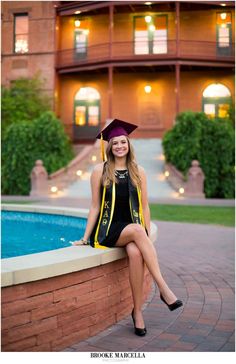 a woman in graduation cap and gown sitting on the edge of a wall next to a pool