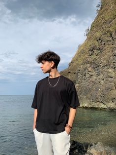 a young man standing on top of a rocky cliff next to the ocean with his hands in his pockets