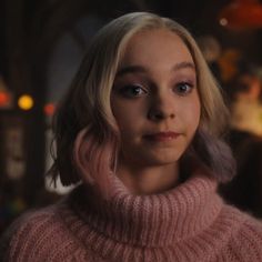 a woman with blonde hair and blue eyes wearing a pink turtle neck sweater in a dimly lit room