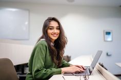 a woman sitting at a desk with a laptop computer in front of her and smiling