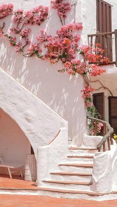 a white building with pink flowers growing on it's side and stairs leading up to the upper floor