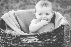 a baby sitting in a basket on the grass with his hand under his chin and smiling