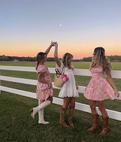 three girls in pink dresses are standing by a white fence and reaching for the moon