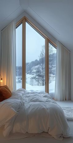 a bed with white sheets and pillows in front of a large window that looks out onto a snowy landscape