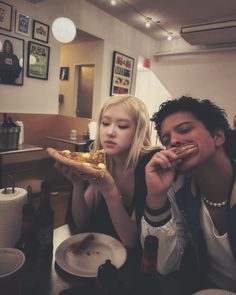 two people sitting at a table with pizza in front of their faces and one person holding a slice of pizza up to her mouth