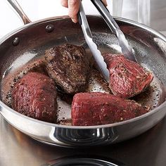 a person is cooking steak in a pan on the stove with tongs and an oven mitt