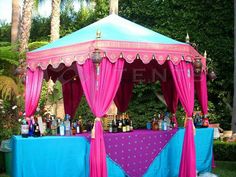 a pink and blue canopy tent with bottles on the table in front of palm trees