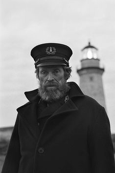 a man with a beard wearing a coat and hat standing in front of a light house