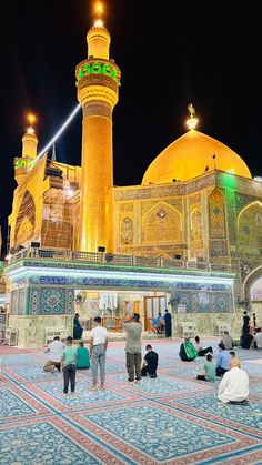 people sitting on the ground in front of a large yellow building with many lights around it