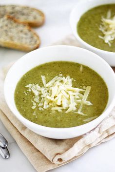 two white bowls filled with green soup on top of a table next to bread slices