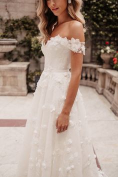 a woman wearing a white wedding dress with flowers on the skirt and off the shoulder