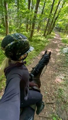 a woman riding on the back of a brown horse down a forest road in front of trees