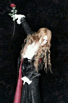 a woman with long hair holding a rose up to her chest and wearing a black dress