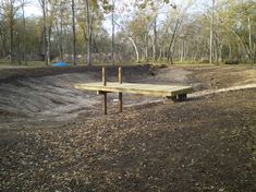 a wooden bench sitting in the middle of a dirt field next to trees and grass