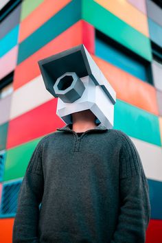 a man wearing a paper mask with a camera on it's head in front of a multi - colored building