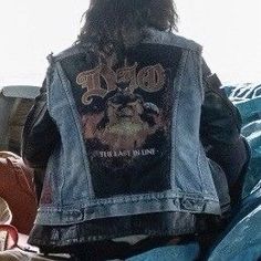 a man with long hair wearing a jean jacket and jeans sitting on top of a bed