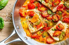 a pan filled with fish and tomatoes on top of a wooden table next to basil leaves