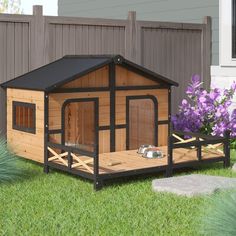 a wooden dog house with a black roof and two dogs in the front yard, next to purple flowers