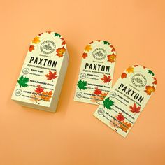 two tags with autumn leaves on them are sitting next to an orange background and one has a white tag that says paxton