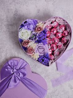 a heart shaped box filled with assorted flowers and chocolates next to a purple bow