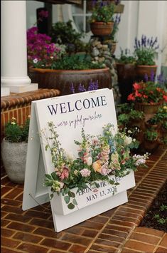 a welcome sign sitting on top of a brick walkway next to potted plants and flowers