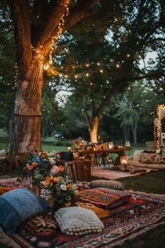 an outdoor seating area is lit up with string lights and pillows on the grass, surrounded by trees