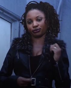 a woman wearing a black jacket and fur collar standing in front of a door with her hand on her hip