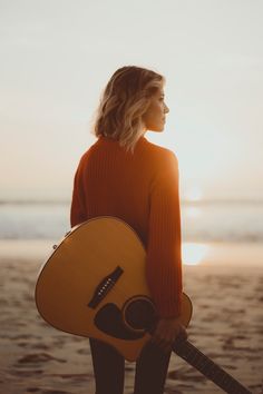 a woman standing on the beach holding a guitar and looking at the sun in the distance