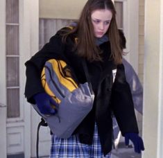 a woman in plaid pants and black jacket carrying a yellow and gray backpack while standing next to a door