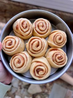 a person holding a metal bowl filled with cinnamon buns on top of a table