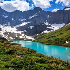 the mountains are covered in snow and green vegetation near a blue lake surrounded by tall grass