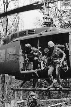 black and white photograph of soldiers climbing into a helicopter in the woods with another man standing next to him