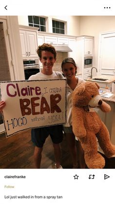 two people standing in a kitchen holding up a sign with a teddy bear on it