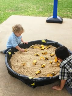 two young boys playing in a sand pit with construction vehicles on the ground next to them