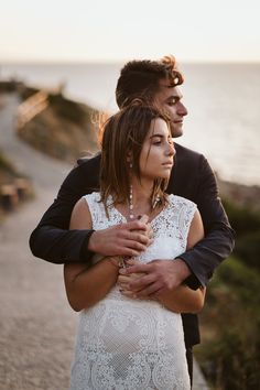 a man and woman standing next to each other in front of the ocean at sunset