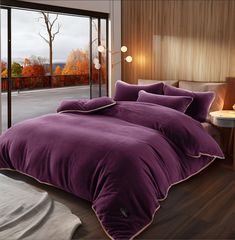 a bed with purple comforter and pillows in front of a large glass window that looks out onto the outdoors