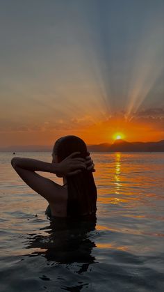 a woman sitting in the water at sunset with her arms around her neck and head