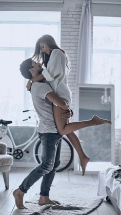 a man holding a woman in his arms while standing on the floor next to a bed