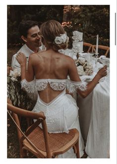 a man and woman sitting at a table with flowers in their hair, dressed in white