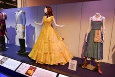 there are many dresses on display in this museum case, and one is wearing a yellow dress